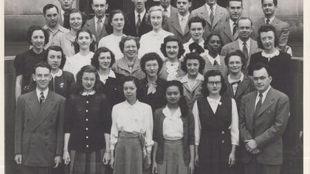 A black and white photo of the class of 1948, wearing skirts and crisp button-downs or suits, with more than 30 people