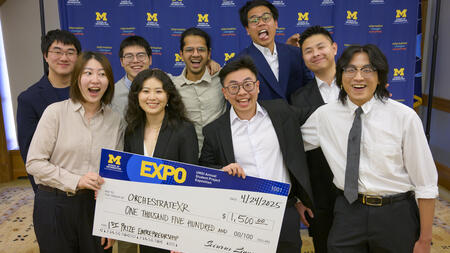 Nine people pose in formal clothes while holding a giant UMS-branded award check