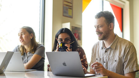 Three students sitting at a table in soft sunlight with laptops in front of them, while one sips from a mug