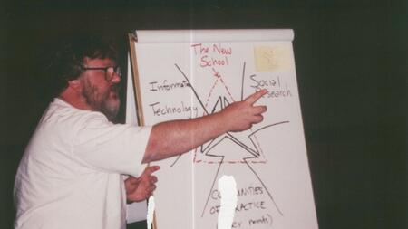 A faculty member gestures in front of a large poster with an illustration that says "The new school" and "Information technology" and "Social research." The photo has some markings of age.