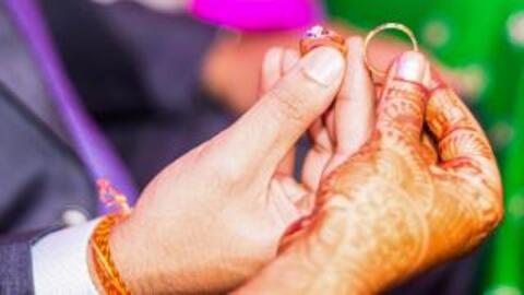 The hands of a bride and groom holding rings. The bride's hand is adorned with intricate henna. 