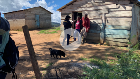 A play button graphic superimposed over a photo of local residents speaking with a Puente volunteer.