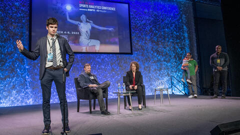 Charlie Logan standing onstage at the MIT Sloan Sports Analytics Conference. Daryl Morey and Jessica Gelman, founders of the conference, sit onstage behind him. 