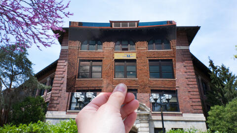 A hand holds up a photo of University of Michigan's West Hall with a School of Information banner hanging from the window in front of the actual West Hall building.