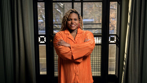 Chelsea Peterson-Salahuddin wearing orange, smiling and standing in front of a window. 