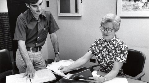 An archival photo of former UMSI professor Constance Rinehart. She is shown sitting at a desk and holding a document. A young man is looking over her shoulder. 