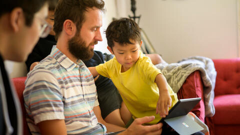 Garret Potter sits on a couch with Max James, holding a laptop. Max reaches toward the laptop screen. Leo looks on from the left side of the frame.