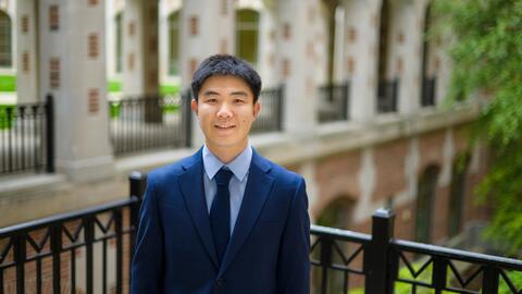 Jiaxin Pei wearing a blue suit and tie, standing outside and smiling at the camera. 