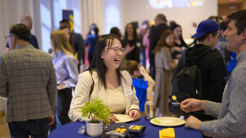 A MADS student laughs at a table while chatting with a faculty member, with a larger reception visible behind them.