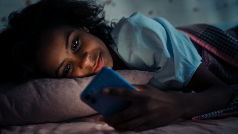 A teenager sitting on her side on a bed smiling at her phone. She's wearing pajamas and it's nigh time. 