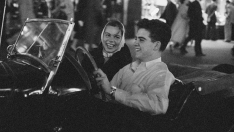 A black and white photo of a teenager driving a classic car while another teen smiles from the passenger seat