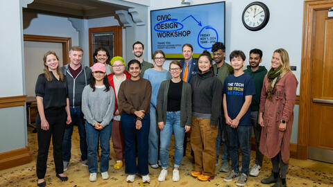 A group of sixteen participants and organizers from the civic workshop pose in front of a lobby monitor that says "Civic Design Workshop"