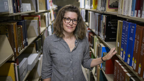 A librarian stands in the aisle between library shelves, with one arm resting on a book