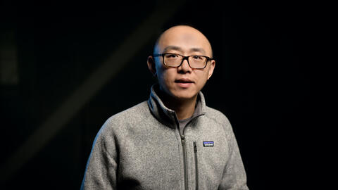 Jiayu Zhou wears a sweater and a black pair of glasses and looks at the camera. 