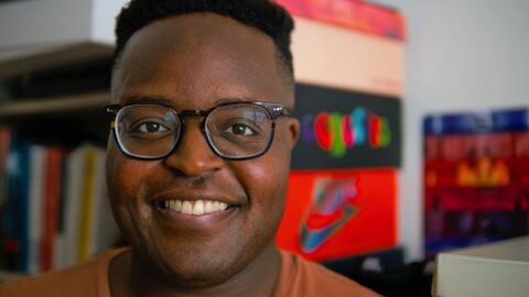 A headshot of Hope Tambala with colorful shelves and file boxes visible behind him