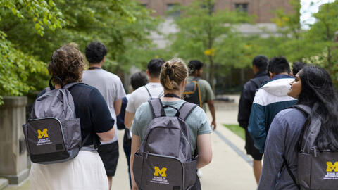 A group of students walk on campus, with three students wearing UMSI backpacks in the foreground 