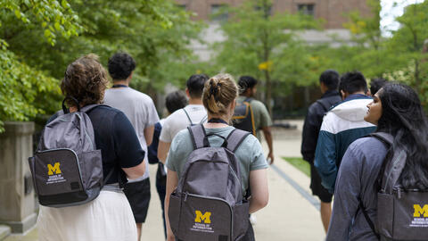 A group of students walk on campus, with three students wearing UMSI backpacks in the foreground 