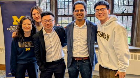 Five students pose, smiling, in a bright classroom setting with a banner behind them that displays the Michigan block M