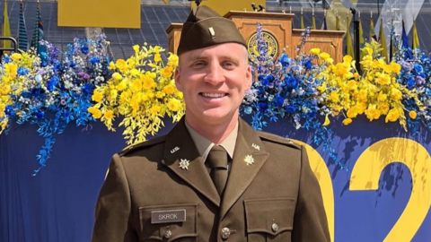 A portrait of Noah Skrok posing in uniform in front of a blue stage decorated with maize and blue floral arrangements, with a podium visible
