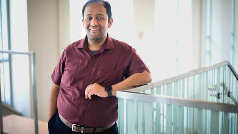 Venkatesh Potluri, a man with short dark hair, wearing a maroon button-up shirt and dark pants, stands indoors leaning casually against a glass railing. He is smiling and has one hand in his pocket, with the other resting on the railing. Soft natural light filters in through large windows behind him.