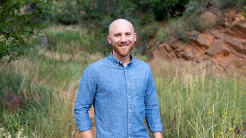Dane Jacobsen smiling outdoors in a grassy landscape featuring craggy rock face.