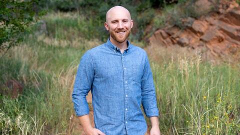 Dane Jacobsen smiling outdoors in a grassy landscape featuring craggy rock face.