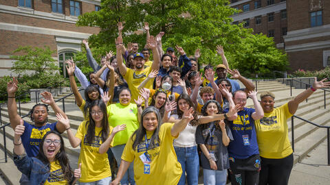 A group of people pose with their arms raised on the steps in front of an academic building