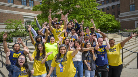 A group of people pose with their arms raised on the steps in front of an academic building