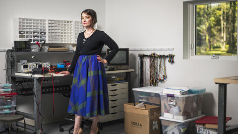 Sophia Bruckner stands in her workshop surrounded by electronics and tools. She wears a black long-sleeve top tucked into a blue and green plaid skirt with tan high heels. One hand rests on a workbench covered with wires, power supplies, and instruments, while the other is on her hip. A window reveals sunlight and trees outside.