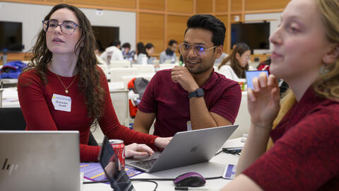 Three teammates, all wearing red, smile and glance to the left. Each has a laptop on a table in front of them in a large academic room