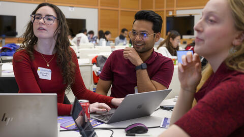 Three teammates, all wearing red, smile and glance to the left. Each has a laptop on a table in front of them in a large academic room