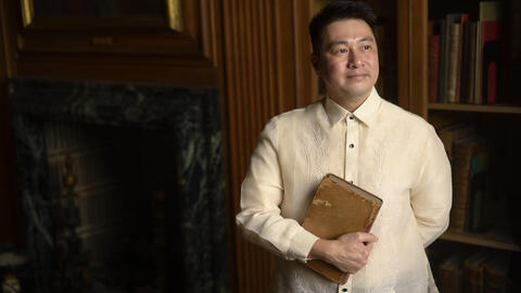 Ricky Punzalan stands in a library in low lighting looking afar and holding a book.  