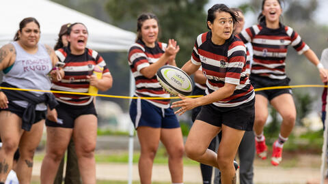 Action shot of Carolann Decasiano holding a rugby ball in the midst of a match, with teammates emoting on the sidelines behind her. They are dressed in matching striped uniforms.