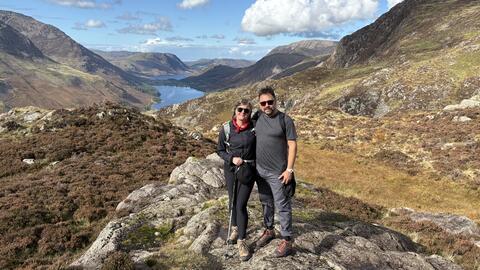 Katie McCurdy stands with another person in a rocky landscape with rolling hills, a body of water and a vast blue sky with billowing clouds stretching far behind them. They are both wearing sunglasses and hiking gear.