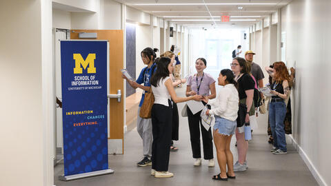 A group of students gather, facing each other and chatting, in a brightly lit hallway with a tall blue UMSI pull-up banner that says Information changes everything