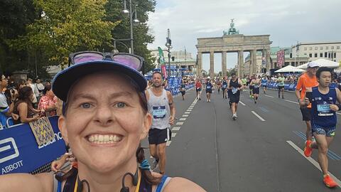 Lydia Schrandt smiling as she takes a selfie while running a race in a city amid other runners wearing race bibs. Onlookers cheer racers on from the sidelines.