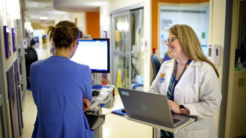 A student stands at a laptop in a hospital hallway smiling while chatting with a provider in blue scrubs