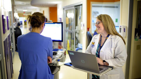 A student stands at a laptop in a hospital hallway smiling while chatting with a provider in blue scrubs