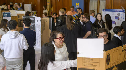 Many people gather in a large room around different posters, with a person gesturing to a poster in the foreground