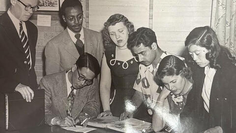 Black-and-white archival photo of seven people gathered around a table, reviewing papers and documents together. One person writes while others stand or sit closely around the materials.