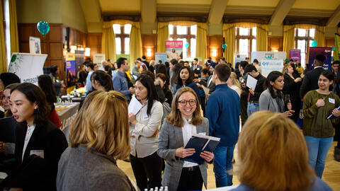 An image of a crowd of students at the UMSI career fair, viewed from a recruiter's perspective behind a table. A particular student is in focus, smiling as they face recruiters.