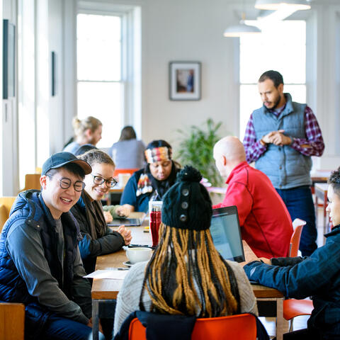 A group of people sit at a table in a light-filled modern room, one on a laptop, their expressions warm and enthusiastic