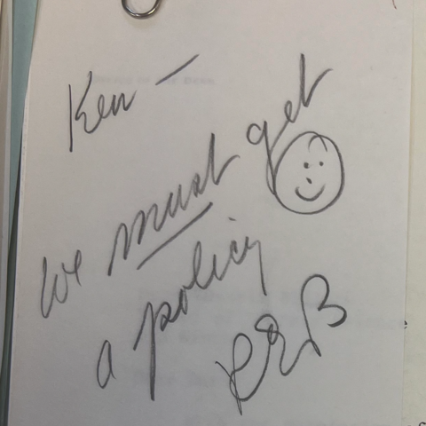A handwritten note that says "Ken, we must get a policy" with a smiley face and the signature of Russel Bidlack