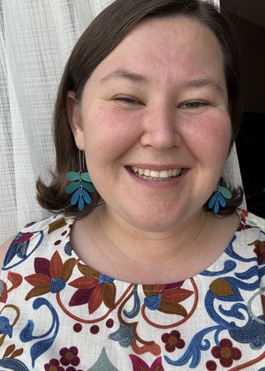 Carina Wilson with short brown hair, blue and green long earrings, and a light tan shirt with pink, blue, purple, and orange flower designs. Standing in front of a white curtain smiling. 