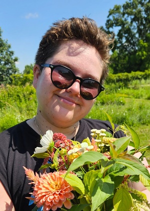Beck smiles while holding a colorful bouquet of flowers. They are a fat white butch wearing sunglasses with cropped hair.