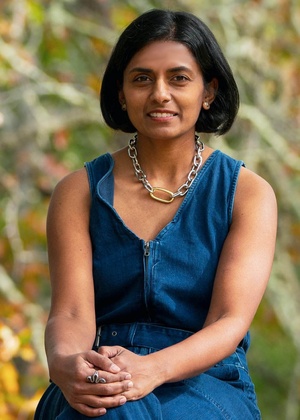 Vidya Setlur seated outdoors, wearing a sleeveless blue dress and silver necklace, against a softly blurred Fall background.