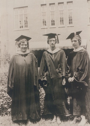 A black and white photo of three young women in caps and gowns, standing in front of a brick building 