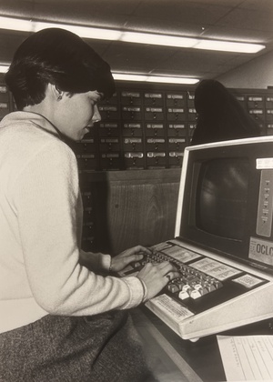 Black and white photo of the side profile of a student typing on a chunky keyboard with card catalogs visible in the background