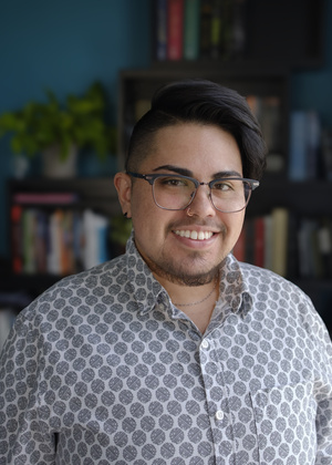 A smiling latiné person with dark swept-back hair, wire glasses, and a short beard, wears a geometric patterned button up shirt, while standing in front of a colorful bookshelf. 