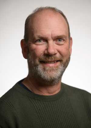 Chris Teplovs smiling at the camera in a dark olive-green crew-neck sweater, with a short grey and reddish-brown beard, against a plain light grey background.
